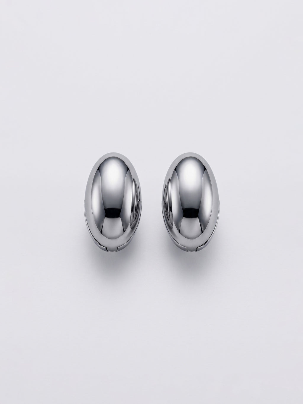 A pair of polished silver-toned metal egg shaped huggie hoop earrings on a clean, light gray background.